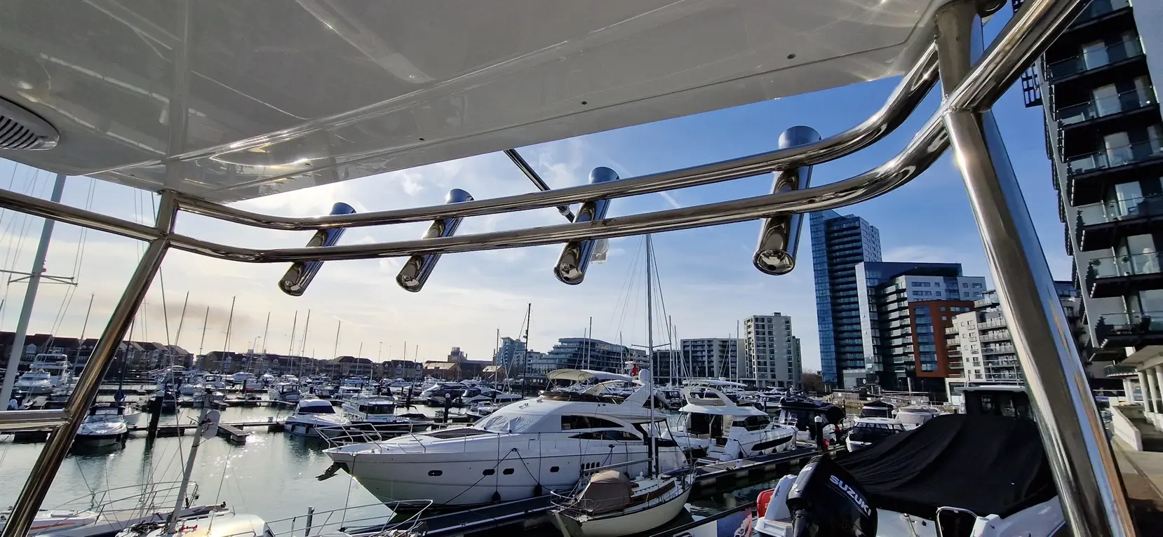 View from a yacht showing the marina with several boats and modern buildings, framed by shiny metal railing.